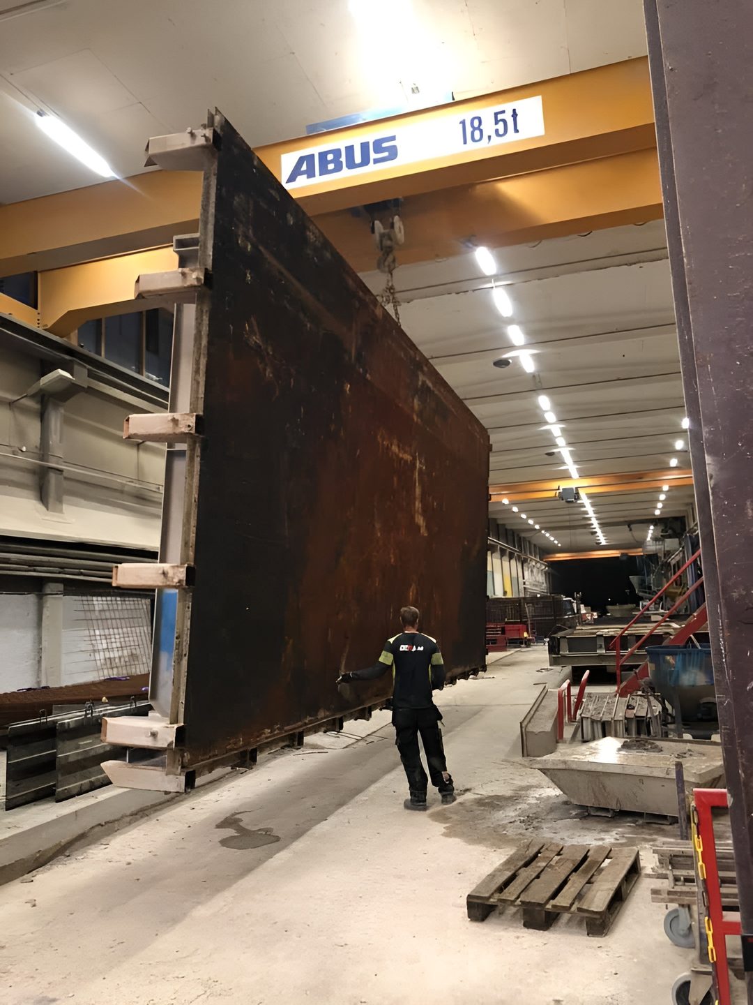 Tekniker från Dema Industripartner AB styr en stor stålform som lyfts med traverskran i en industrihall.