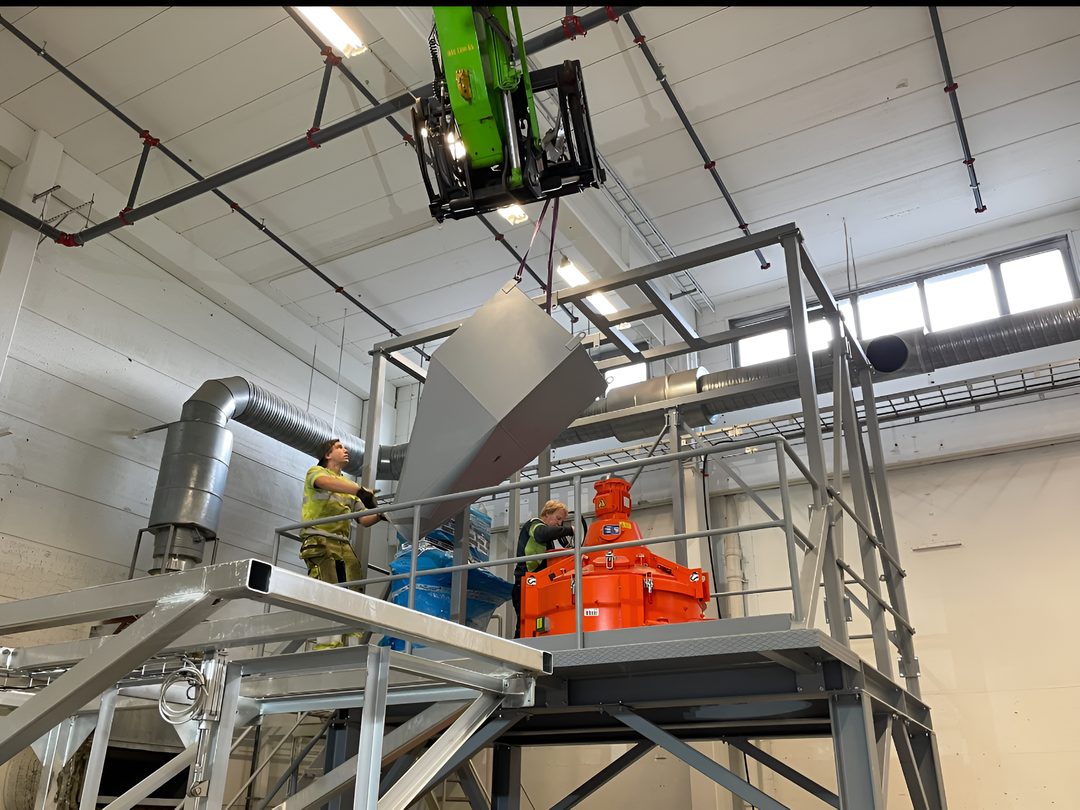 Två tekniker från Dema Industripartner AB monterar en stålkanal som lyfts på plats av en lyftkran i en industrihall.