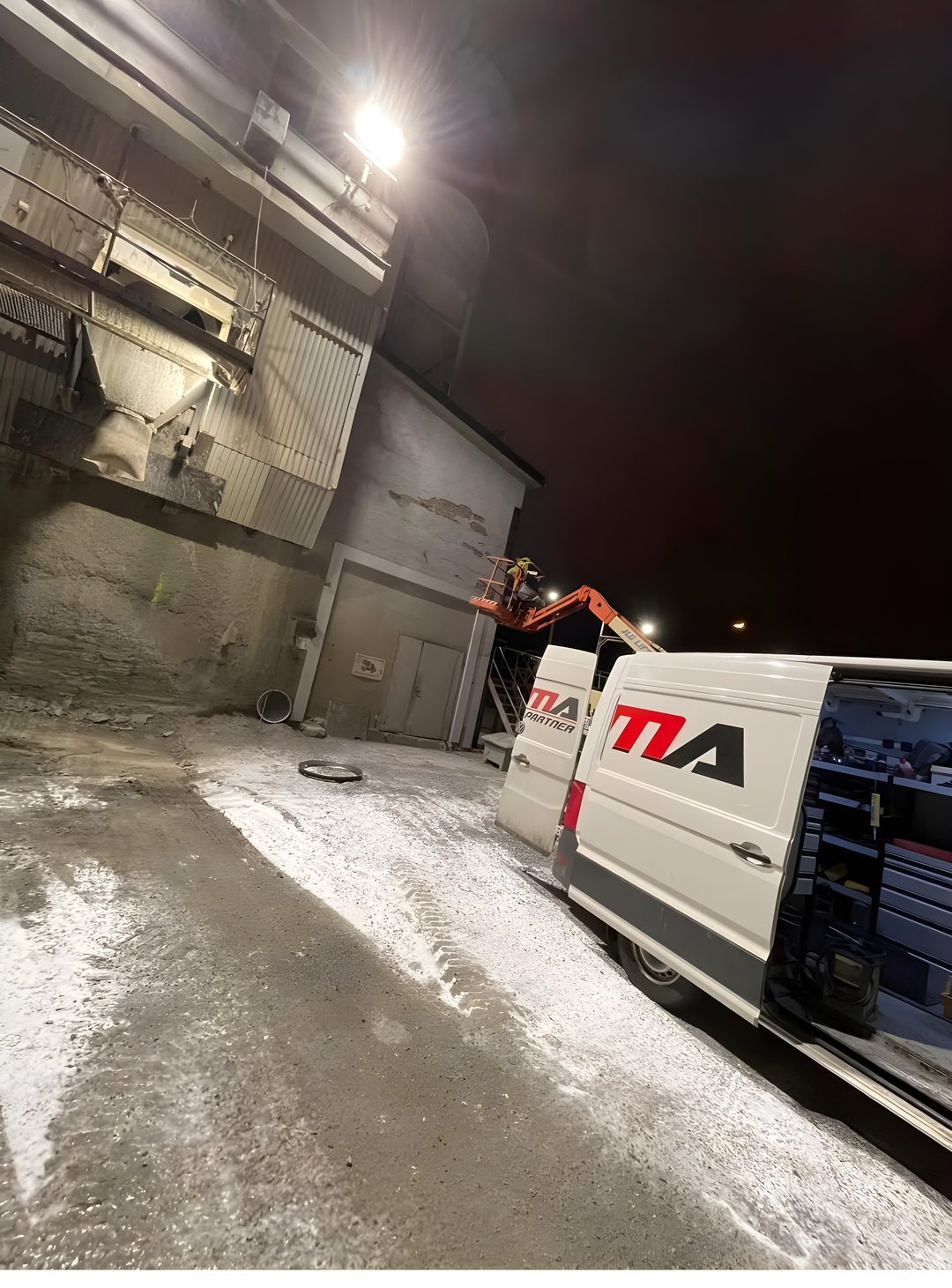 Demas servicebil och tekniker i skylift som utför servicearbete på en industrianläggning under vinternatt.