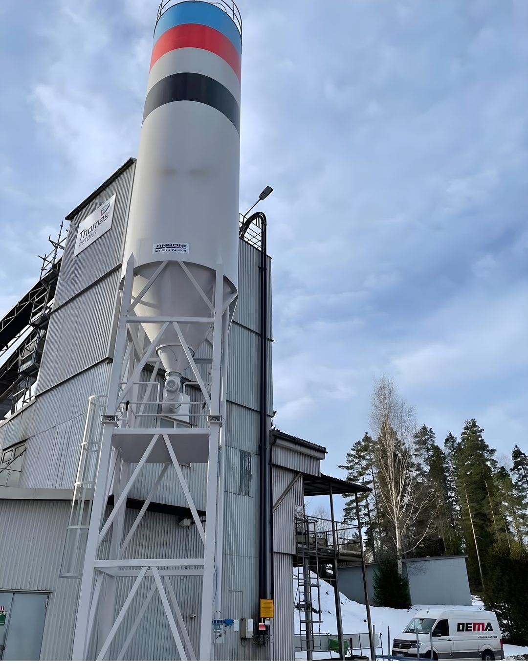 Vit industrisilo med röd, blå och svart märkning vid en betongstation, vintermiljö med Demas servicebil.